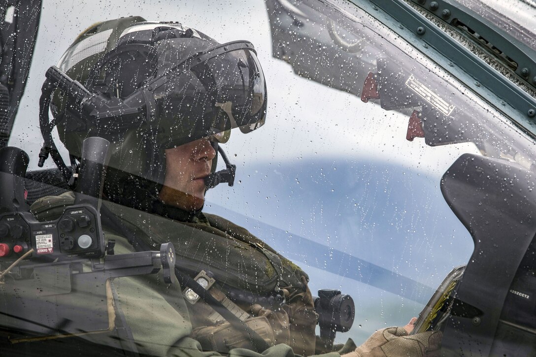 A pilot sits in a helicopter cockpit with water droplets on the windshield.