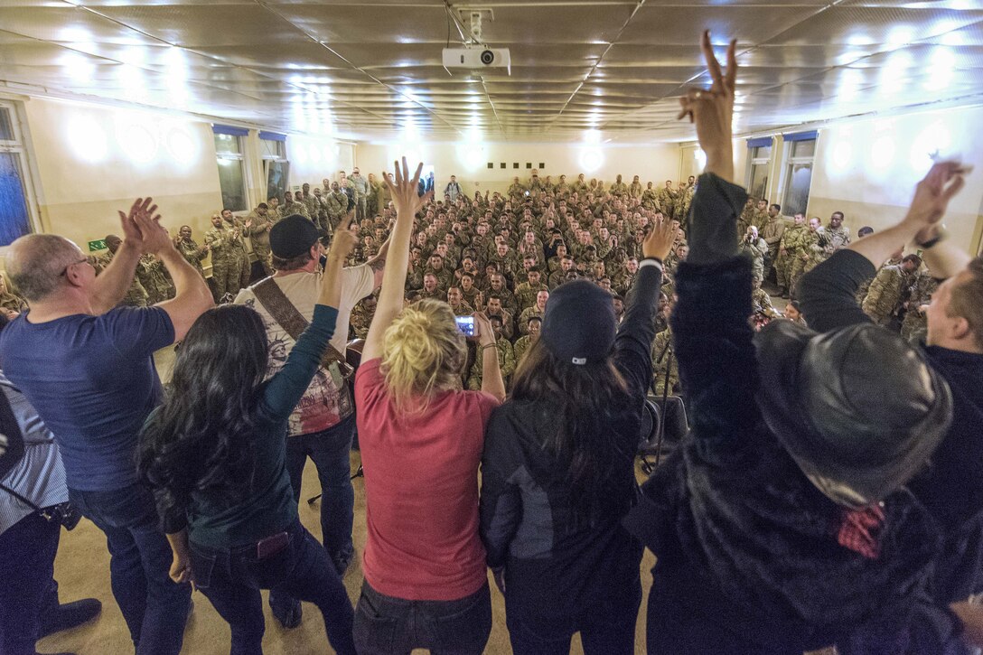 An audience of troops applaud as performers on stage, shown from behind, wave, applaud and gesture.