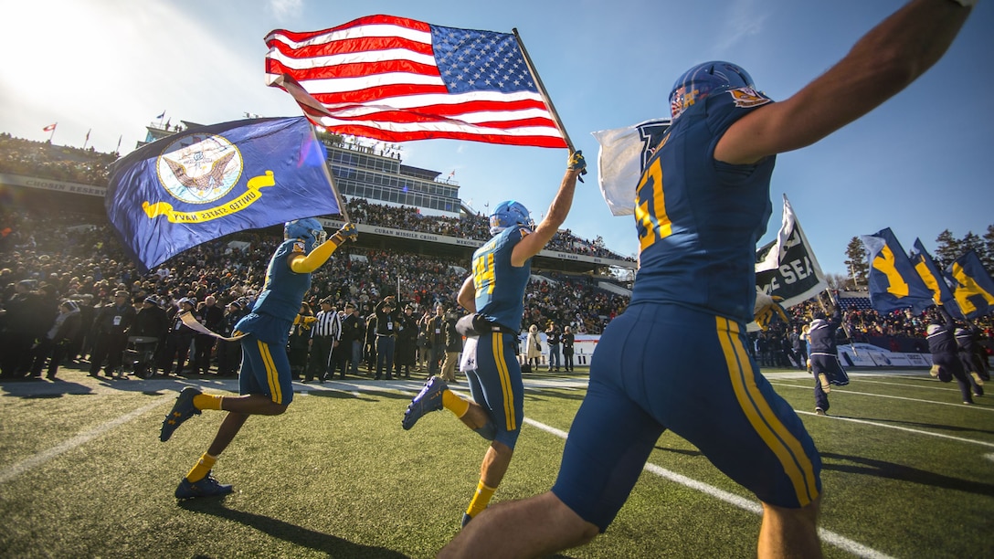 Navy Midshipmen run onto the football field holding flags.