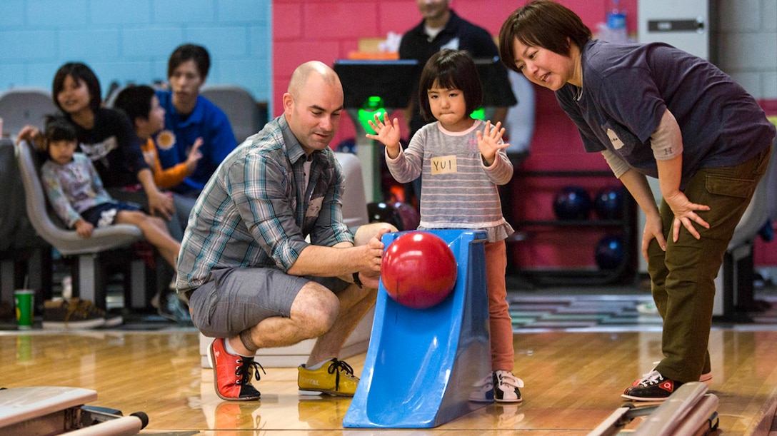 A child rolls a bowling ball down a ramp with two adults nearby.