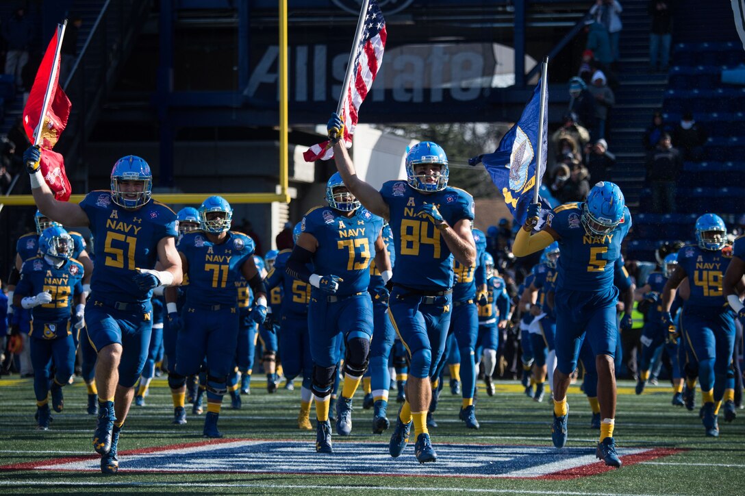 Navy football players, some holding flags, run onto the field.