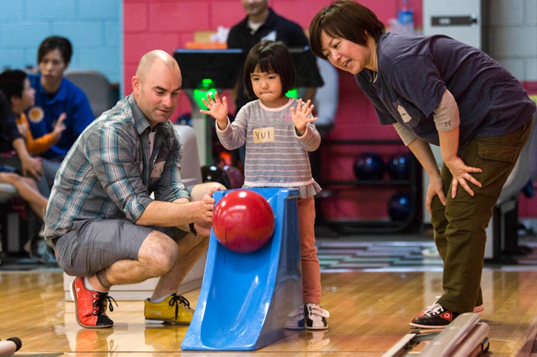 A child rolls a bowling ball down a ramp with two adults nearby.