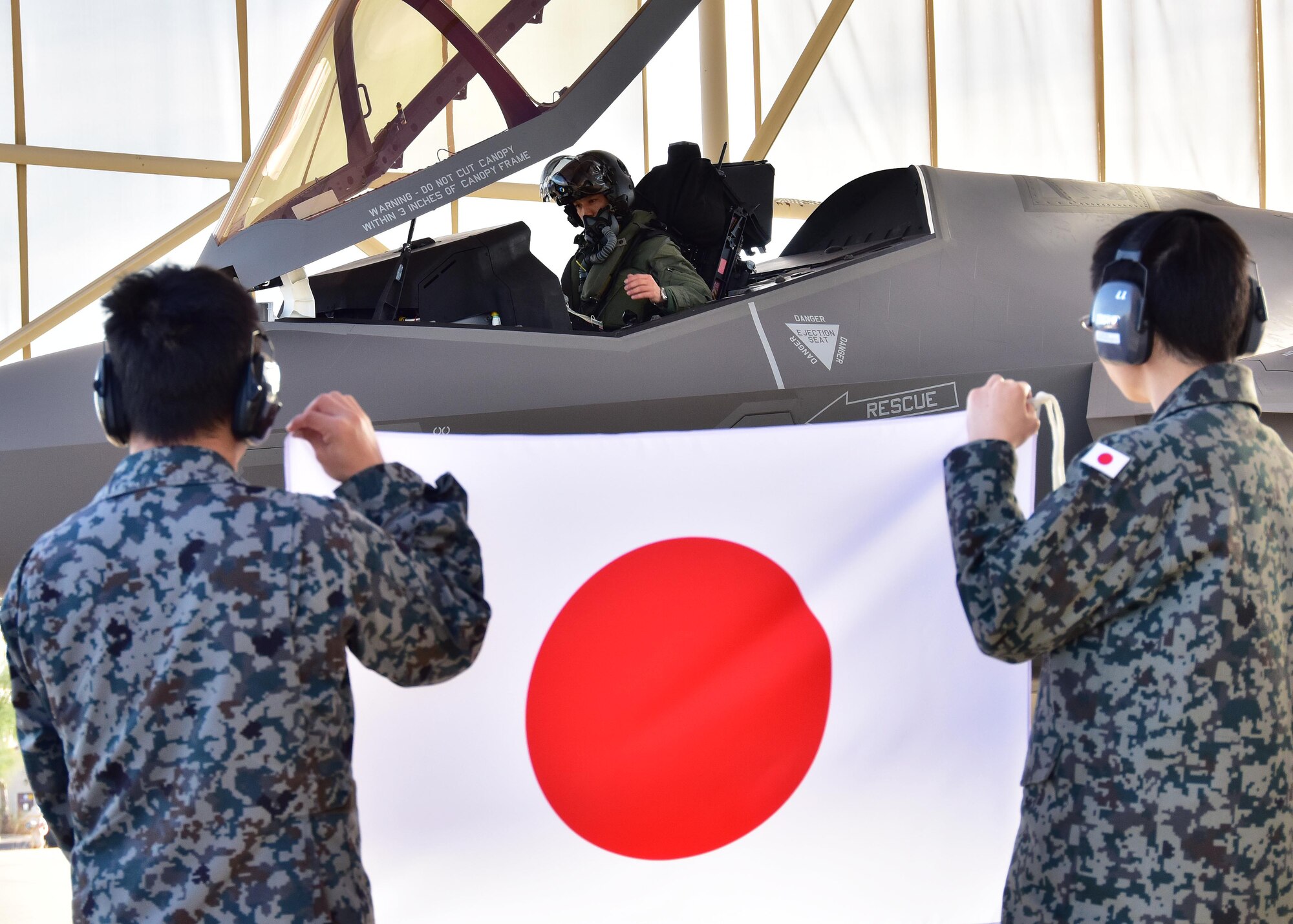 Lt. Col. Nakano, Japan Air Self-Defense Force F-35A national representative pilot to Luke, sits in the cockpit of one of three F-35s on 944th Fighter Wing’s ramp area Feb. 7 before departing while two JASDF maintainers hold a Japan flag up at Luke Air Force Base, Ariz. (U.S. Air Force photo taken by Tech. Sgt. Louis Vega Jr.)