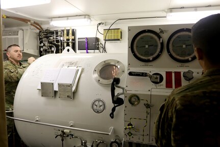 Master Diver 1st Sgt. Tyler Dodd, with the 511th Engineer Dive Detachment out of Fort Eustis, Va., oversees training of the Standard Navy Double Lock Recompression Chamber System while salvage diver Spc. Nathaniel Marquez works the control console, Kuwait Naval Base, Kuwait, Feb. 13, 2017. The training is to give 511th Soldiers hands on training in equipment operation, Soldier advancement and to help a Soldier with an injury sustained during a previous training exercise.