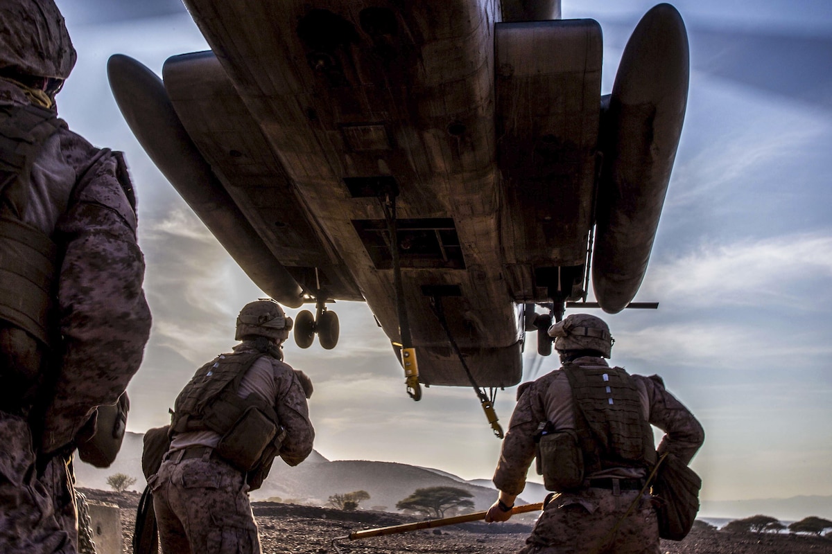Marines stand under a hovering helicopter as they prepare to attach a barrier to it.