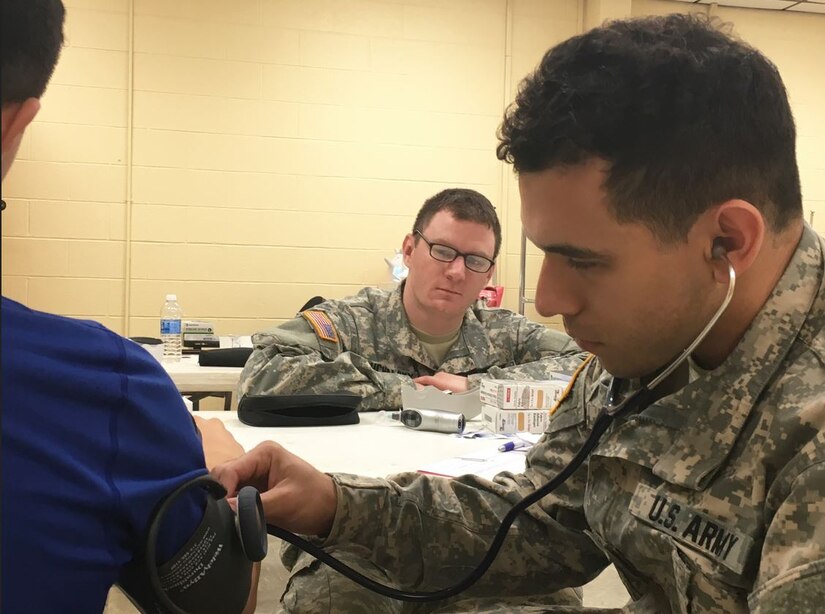 Army Reserve Pfc. Stephen Young, right, checks vitals on a patient as Sgt. Kyle Puchalsky, center, observes during Operation Lone Star at Vela Middle School in Brownsville, Texas, July 25, 2016. Both Puchalsky and Young are health care specialists with the 399th Combat Support Hospital. This year, Army Reserve medical professionals will partner with Texas A&M nurse practitioner students to provide medical care at four sites in colonias near Laredo, Texas, June 17 to July 1. (Photo Credit: Staff Sgt. Syreeta Shaw)