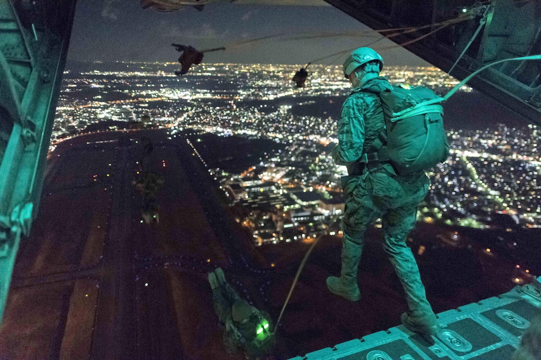 A Marine, shown from behind, steps off the back of an open aircraft into the night sky.