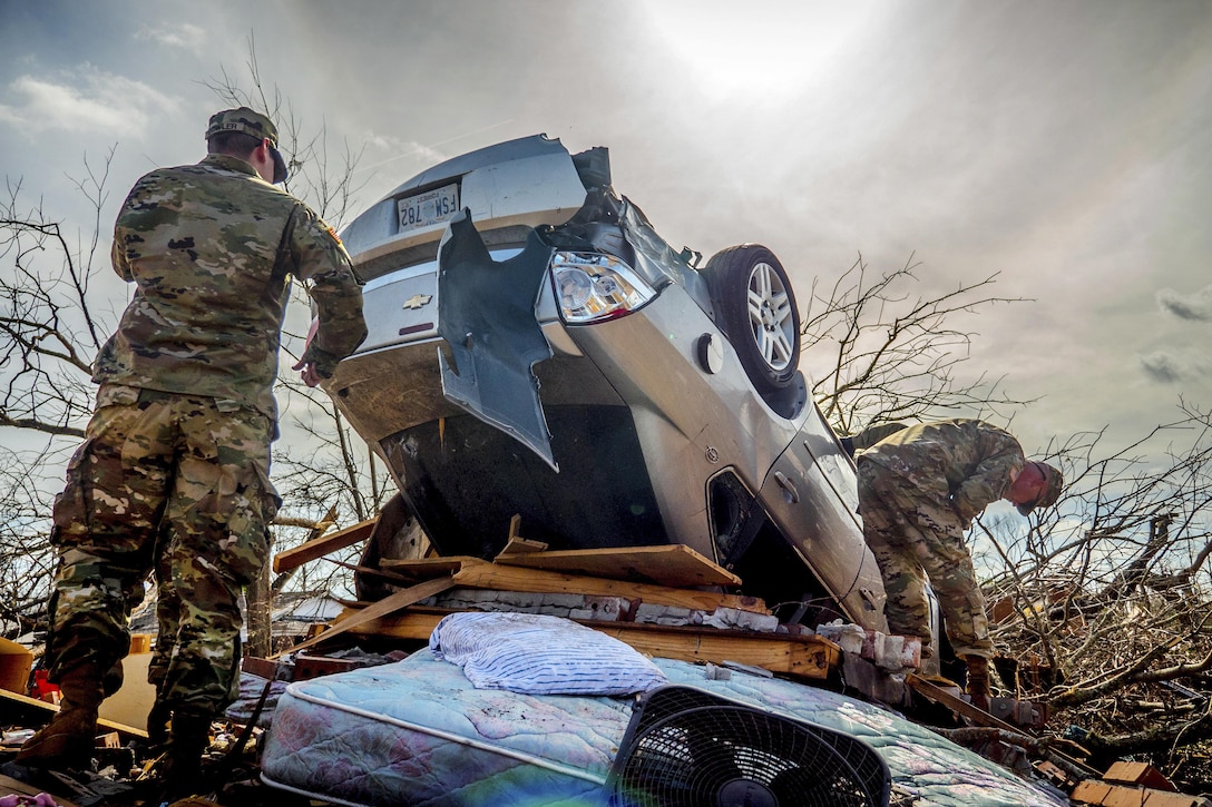 Soldiers inspect a car that was flipped over by a tornado and sits on top of a pile of debris.