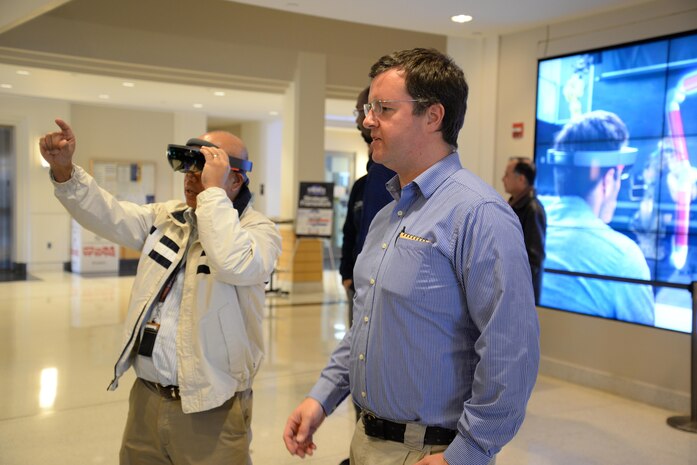 Joseph Curran, an ocean engineer with the Welding, Processing and Nondestrcutive Evaluation Branch at Naval Surface Warfare Center, Carderock Division, explains how to use the HoloLens to Henry Molintas, a mechanical engineer in Carderock's Propulsor Manufacturing Office. The Microsoft HoloLens is an augmented reality device that allows the user to still see the real world with an overlay of something else, like a schematic. The technology may be useful to the Navy in areas such as training and maintenance. (U.S. Navy photo by Kelley Stirling/Released)