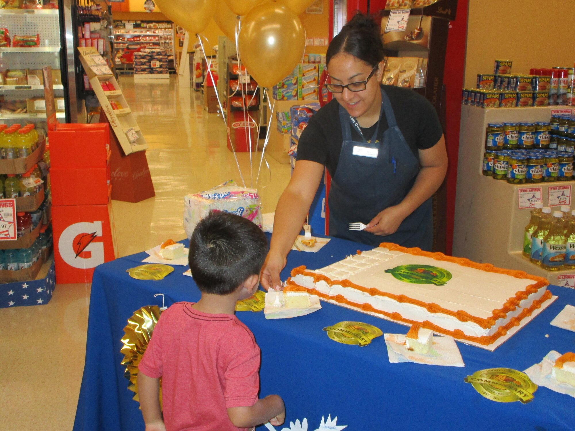 Wignelia Cruz-Gonzalez, Laughlin Commissary produce manager, gives a slice of cake to a military child at Laughlin Air Force Base, Tx., July 1. To observe the commissary’s 150th birthday, Laughlin’s commissary staff handed out free cake and reduced its store prices on food and household goods. 