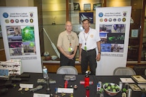 Mr. Paul Burke, Joint Non-Lethal Weapons Directorate Combatant Command (COCOM) Liaison Officer to U.S. Special Operations Command (left), and Mr. Rick Bartis, COCOM Liaison Officer to U.S. Central Command (CENTCOM), stand behind the non-lethal weapons and munitions display at the U.S. CENTCOM Family Day. Nearly 2,500 family members attended the June 16 event.
