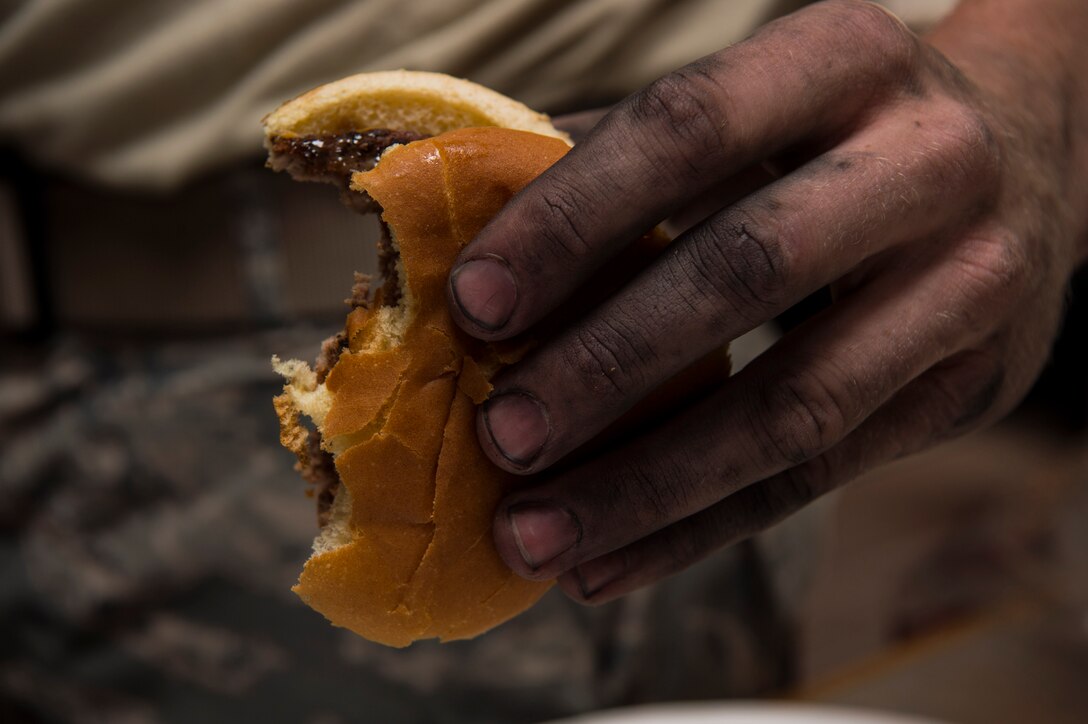The machine-stained hand of Senior Airman Gabriel Chrum, 90th Missile Maintenance Squadron electro mechanical team technician, grasps a hamburger during the 319th Missile Squadron code change burger burn in the 90th Missile Wing missile complex, July 12, 2017. The different units responsible for cleaning sensitive components in the missile complex were offered a freshly-cooked meal by leaders in the 319th Missile Squadron and 90th MW chaplains. (U.S. Air Force photo by Staff Sgt. Christopher Ruano)

