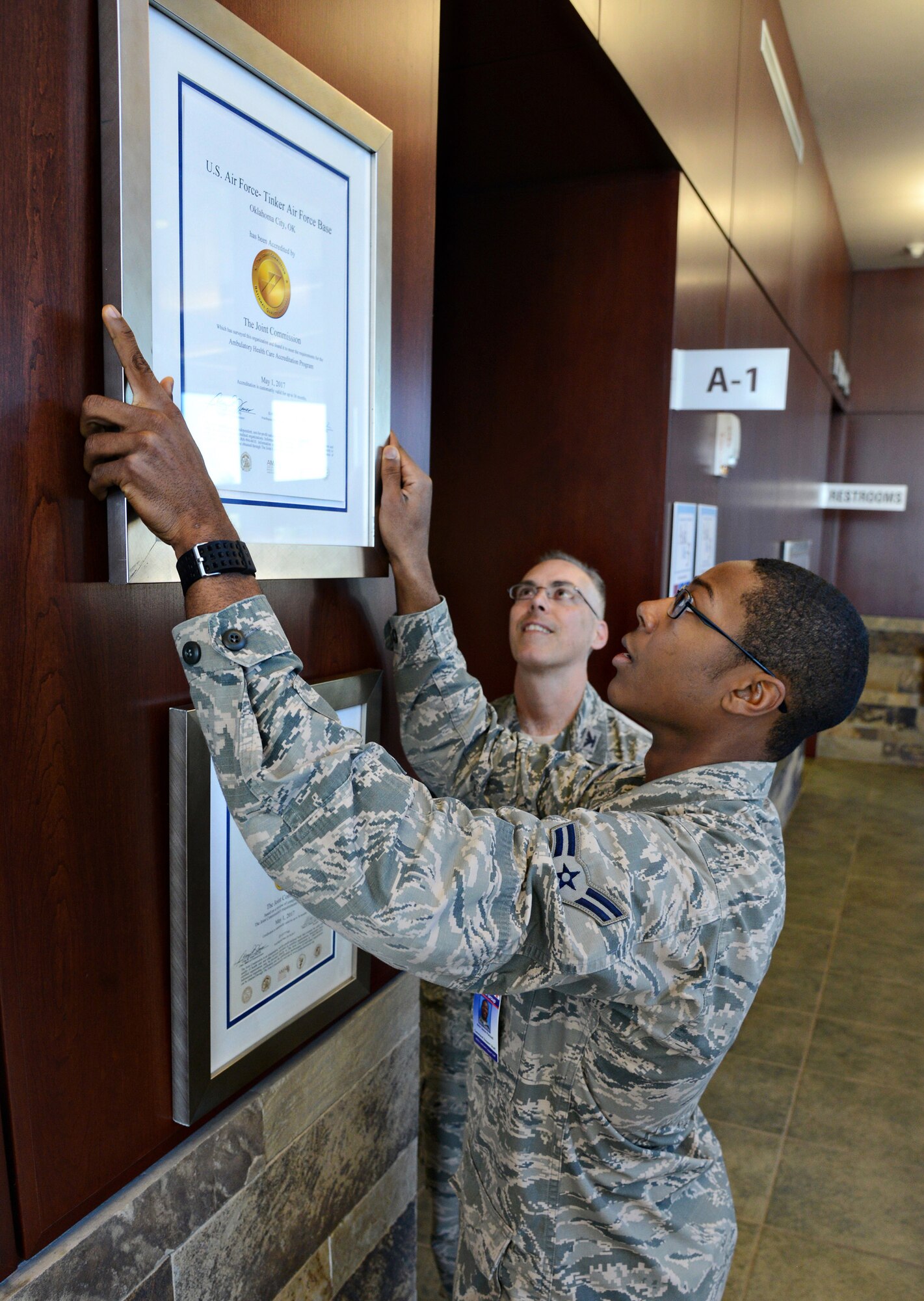 While 72nd Medical Group Commander Col. Christopher Grussendorf watches, Airman 1st Class Jason Pannell, with the 72nd Medical Support Squadron, carefully hangs a certificate from The Joint Commission, bestowing a three-year accreditation to the 72nd MDG by meeting the requirements for the Ambulatory Health Care Accreditation Program.