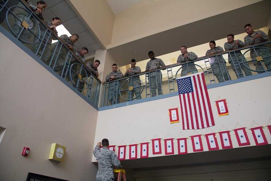 A member of the 66th Security Forces Squadron hangs a service banner July 18 in the atrium of Building 1725 as part the squadron’s tradition for troops who are deploying, while SFS Airmen look on. Members of the squadron recently embarked on an overseas deployment. (U.S. Air Force photo by Mark Herlihy)