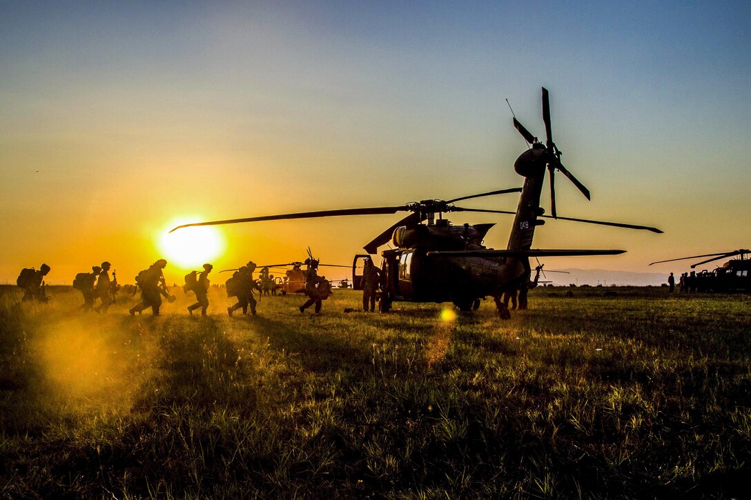 A line of soldiers, shown in silhouette, board a helicopter, with a low sun lighting the sky in the background.