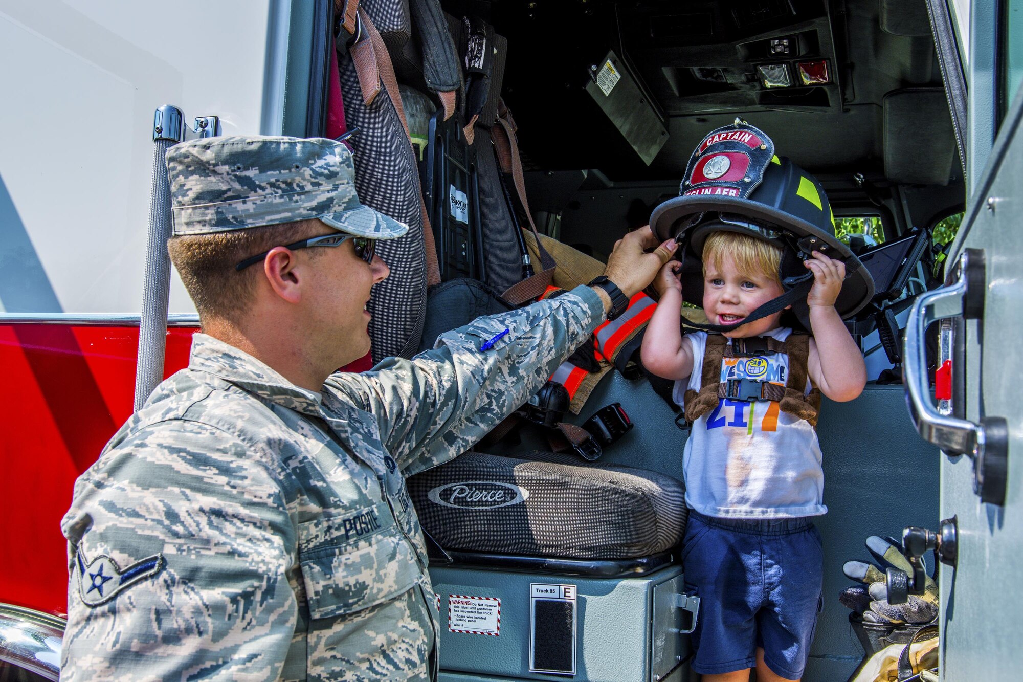 Airman Basic Bryce Postle, 445th Airlift Wing journeyman firefighter, helps hold a firefighter helmet for a toddler during Big Truck Day, July 28, Destin, Fla. Fire, construction, garbage and utility trucks were on display for kids to see up close at the city’s annual event. (U.S. Air Force photo/Kristin Stewart)