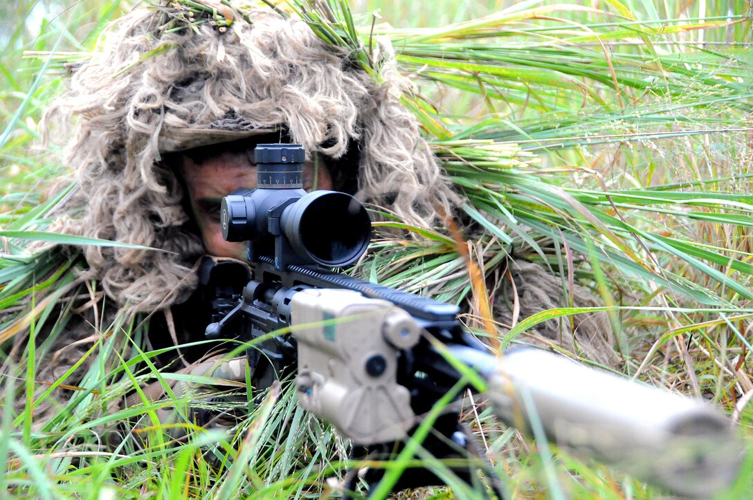 A sniper wearing camouflage and pointing a weapon lays on the grass.