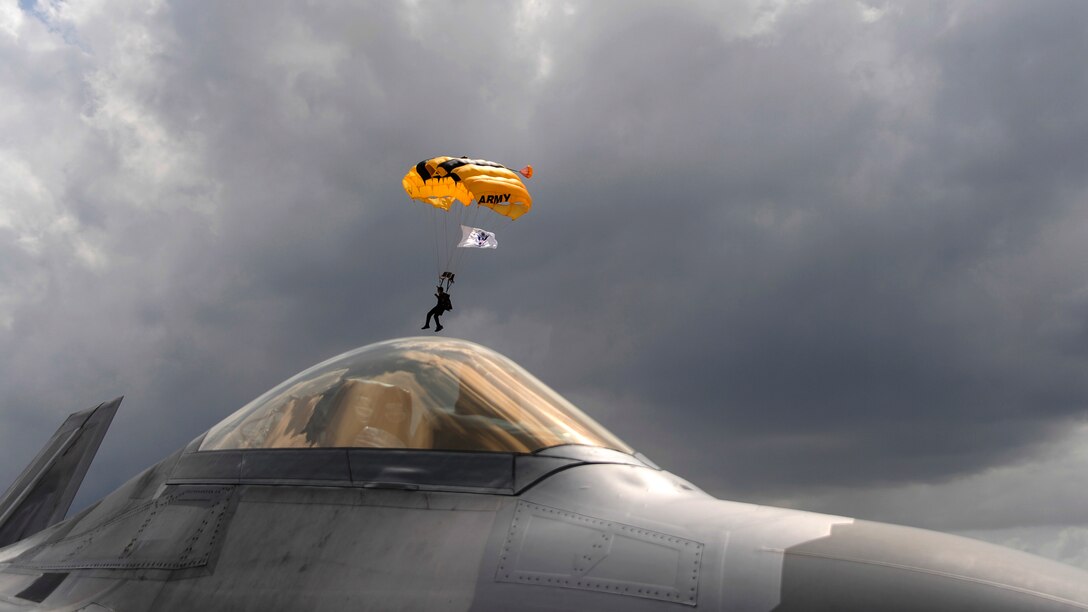 An Army Golden Knights parachute team member descends in the distance as an Air Force F-22 Raptor sits parked at Coast Guard Air Station Miami in Opa-locka, Fla., May 26, 2017, during the National Salute to America’s Heroes Air and Sea Show. Air Force, Army, Navy, Marine Corps and Coast Guard service members participated in the event. Air Force photo by Senior Airman Erin Trower