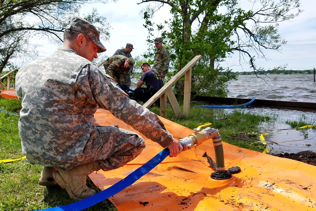 A soldier uses a hose to put water in an orange structure on the ground by a waterway.