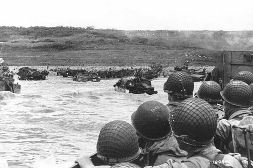 Soldiers in helmets look out at a beach from a landing craft.
