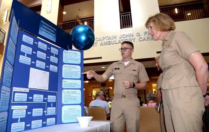 U.S. Navy Ensign Matthew Evans, officer in training for submarine warfare qualification, serving as support staff for the pharmacy at Naval Health Clinic Charleston, shows NHCC Commanding Officer, Capt. Elizabeth Maley, the pharmacy's new procedure for filling prescriptions during Naval Health Clinic Charleston’s 2nd Continuous Performance Improvement Fair here, June 2. The procedure minimizes errors while improving wait times. Evan's display was one of many at the CPI fair designed to highlight the strides taken by NHCC staff members to improve the quality, education and safety of health care for NHCC's patients.