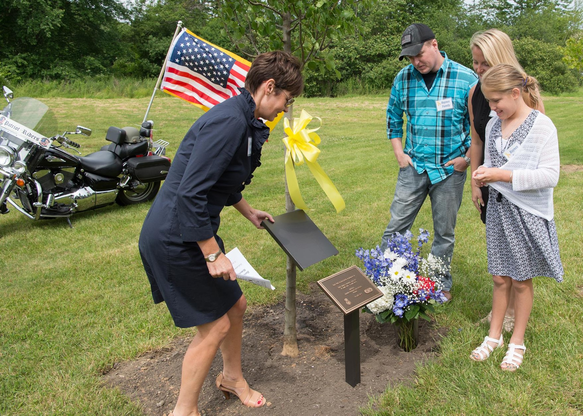 Shelly Raynes unveils a plaque in front of a tree dedicated to her late husband, retired Tech. Sgt. John A Raynes, during a Memorial Park Dedication Ceremony at Hanscom Air Force Base, Mass., June 16, while John Jr., Ashley and Hannah Grace look on. Raynes worked for Security Forces, Public Affairs and Civil Engineering during his tenure at Hanscom. Also honored during the event was Dana E. Kirane and Dennis “Dennie” Guthrie. (U.S. Air Force photo by Jerry Saslav)
