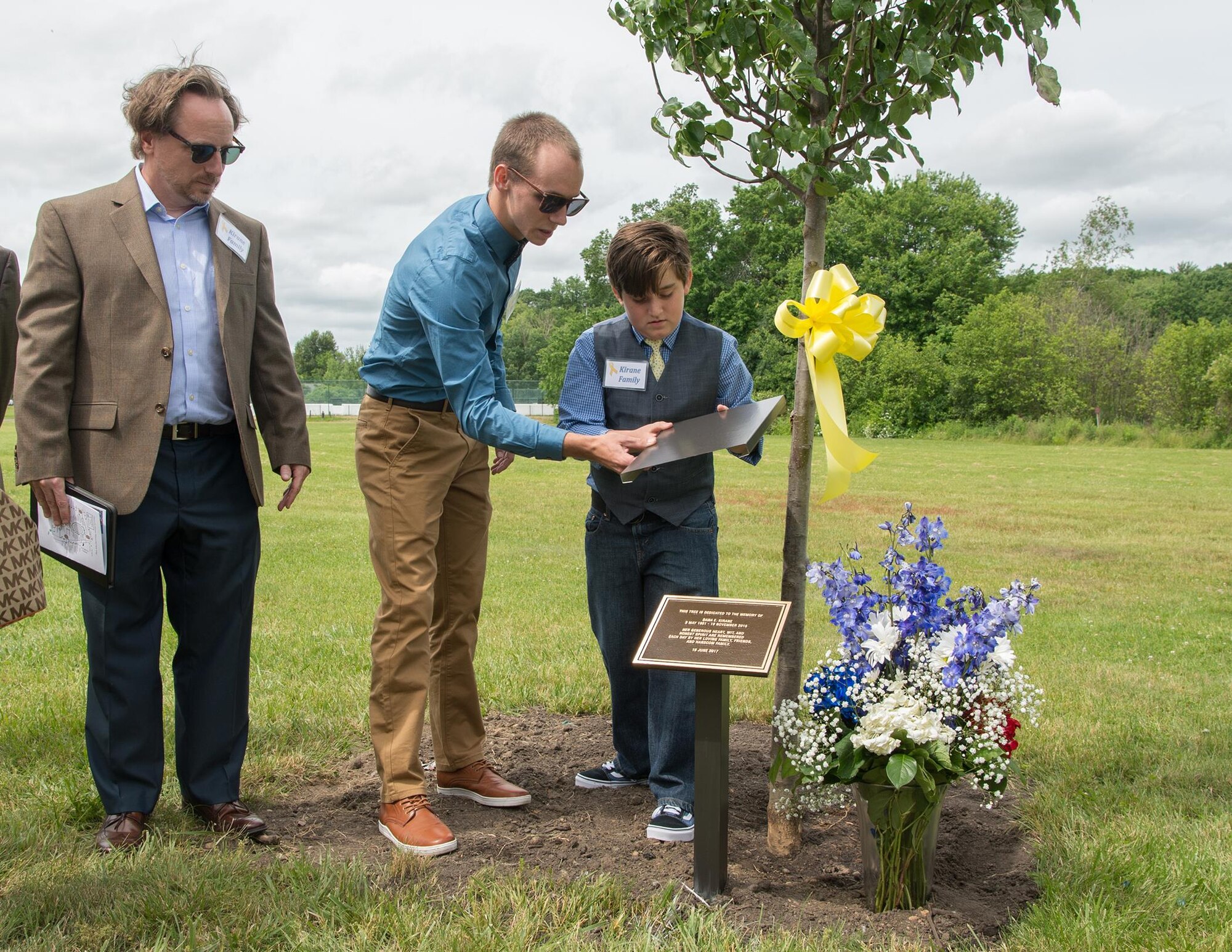 Joe Kirane look on as his children, Dylen and Ian, unveil a plaque in front of a tree dedicated to his mother, Dana E. Kirane, during a Memorial Park Dedication Ceremony at Hanscom Air Force Base, Mass., June 16. Kirane worked at Hanscom from 1974 until her retirement in 2006 as a financial services officer. Also honored during the event were Dennis “Dennie” Guthrie and retired Tech. Sgt. John A. Raynes. (U.S. Air Force photo by Jerry Saslav)