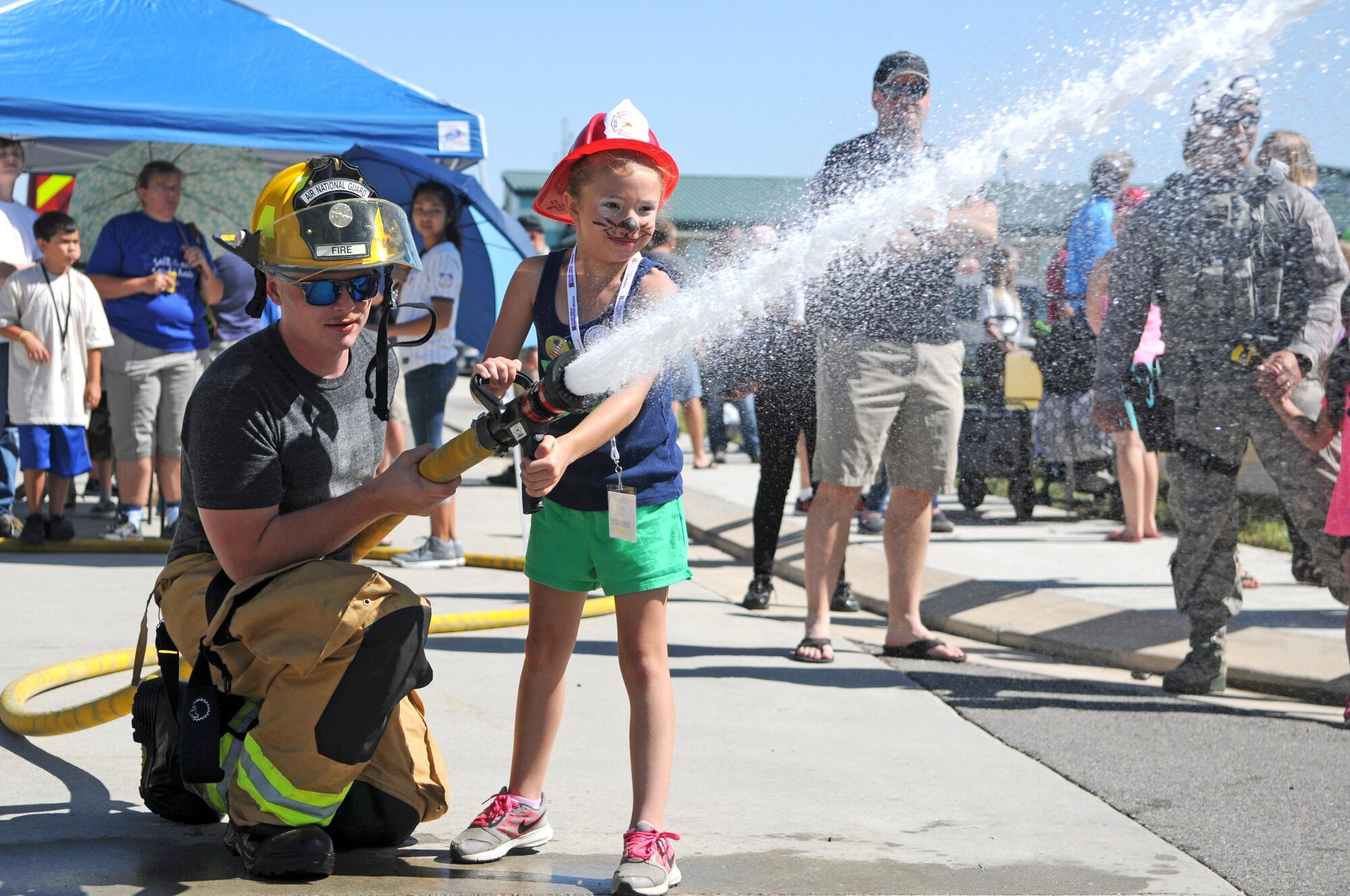 Senior Airman Brody Sanzone, a firefighter with the 151st Civil Engineering Squadron, helps a young girl direct a fire hose toward an orange traffic cone to simulate extinguishing a fire during Wingman Day on June 10, 2017 at Roland R. Wright Air National Guard Base in Salt Lake City. (U.S. Air National Guard photo by Tech. Sgt. Amber Monio)