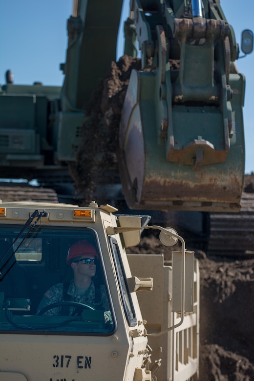A soldier from the 317th Engineer Company watches his mirrors as a Hydraulic Excavator (HYEX) swings a bucket full of dirt over the back of his M1157 A1P2 10-Ton dump truck. The soldier is taking part in the Blackfeet Innovative Readiness Training (IRT) project. The IRT project produces military readiness while simultaneously providing quality services to communities throughout the U.S. This particular project takes place on the Blackfeet Reservation in Browning, Montana during the months of June thru August (U.S. Army photo by Staff Sgt. Jason Proseus/released).