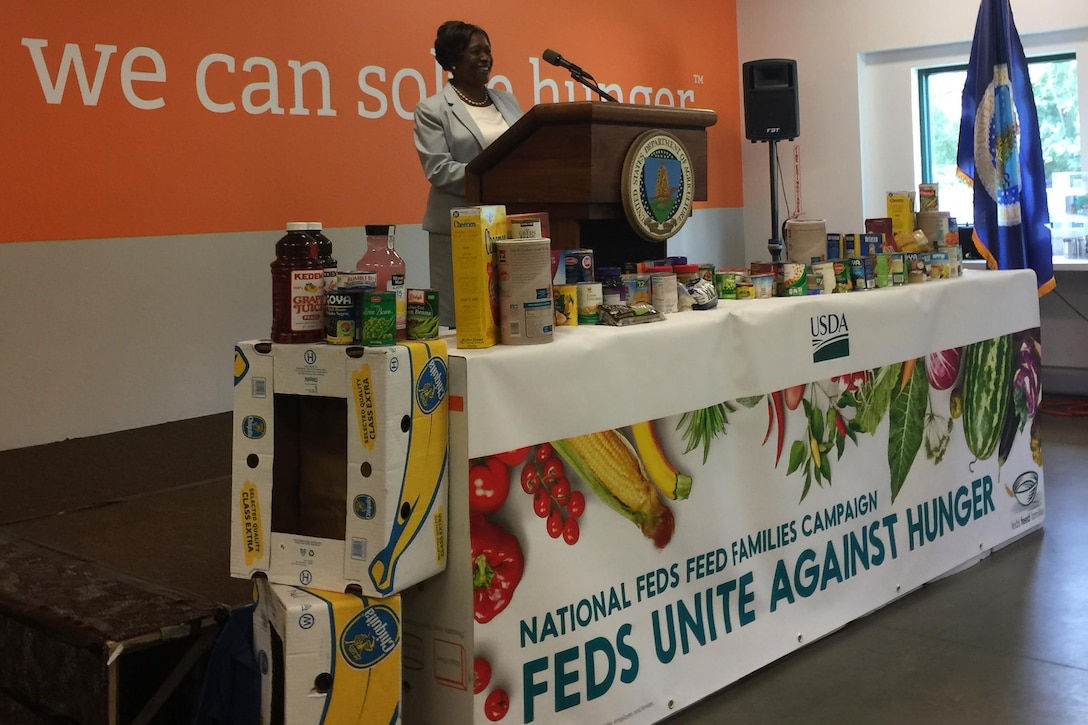 This image shows a woman speaking at a podium behind a table of nonperishable food items.