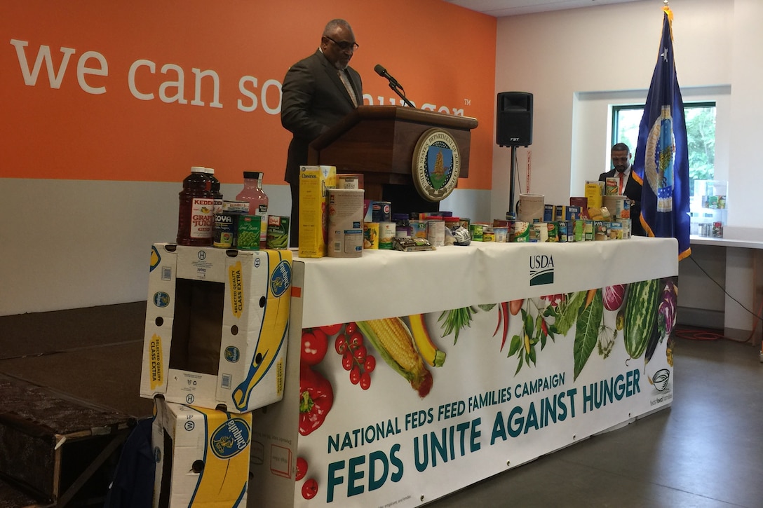 This image shows a man speaking at a podium behind a table of nonperishable food items.