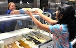 Debra Bratton hands Kartha Navya a plate during lunch in the Brooke Army Medical Center dining room June 20. BAMC Culinary and Hospitality Branch would like patrons to ask for a regular plate rather than a to-go container when eating in the dining facility. 