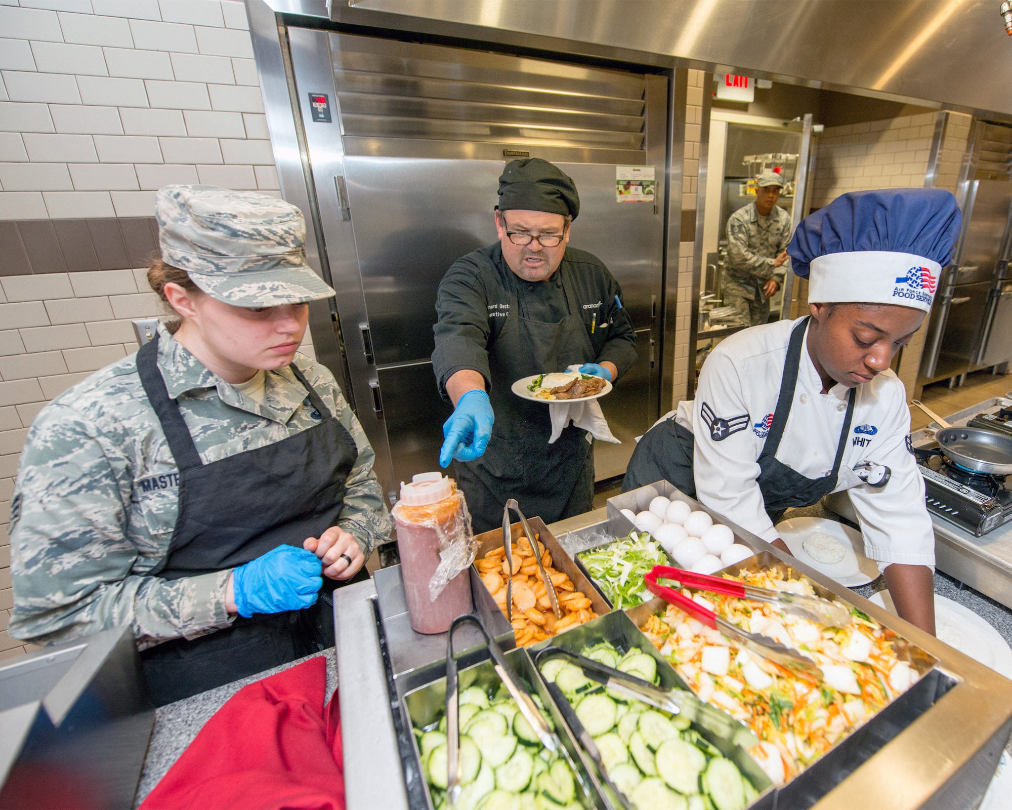 U.S. Air Force Senior Airman Kylee Masters Thomas (left), Maynard Oestreich, executive chef for Aramark, and Airman 1st Class Cierra White (Right), prepare meals prior to the BIBIM Box tasting featuring Korean food at Sierra Inn Dining Facility, Travis Air Force Base, Calif., June 29, 2017. Oestreich a former U.S. Navy veteran and an award-winning chef from Napa Valley, Calif., took the head chef position so he could mentor young Airmen. (U.S. Air Force photo by Louis Briscese)