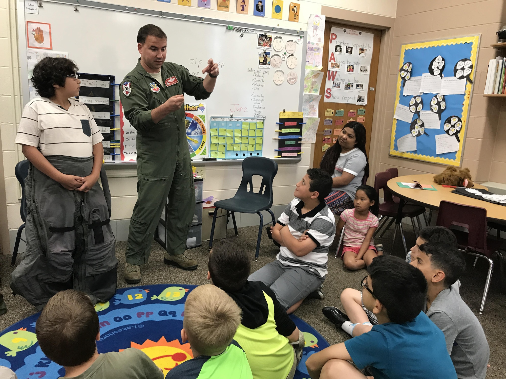 Col. Tom Bladen, 104th Fighter Wing operations group commander, visits refugee students at Highland Elementary School in Westfield, Massachusetts, June 26, 2017. The visit served as a means to help the children understand why they hear the loud screeching F15 jets flying overhead when playing outside and to help them understand the 104th Fighter Wing flies to protect. Bladen explains how the flight suit prevents the pilots from passing out while flying as he speaks to the students after helping the honoree pilot of the day dawn a 104th Fighter Wing flight suit. (U.S. Air National Guard photo by Senior Master Sgt. Julie Avey)