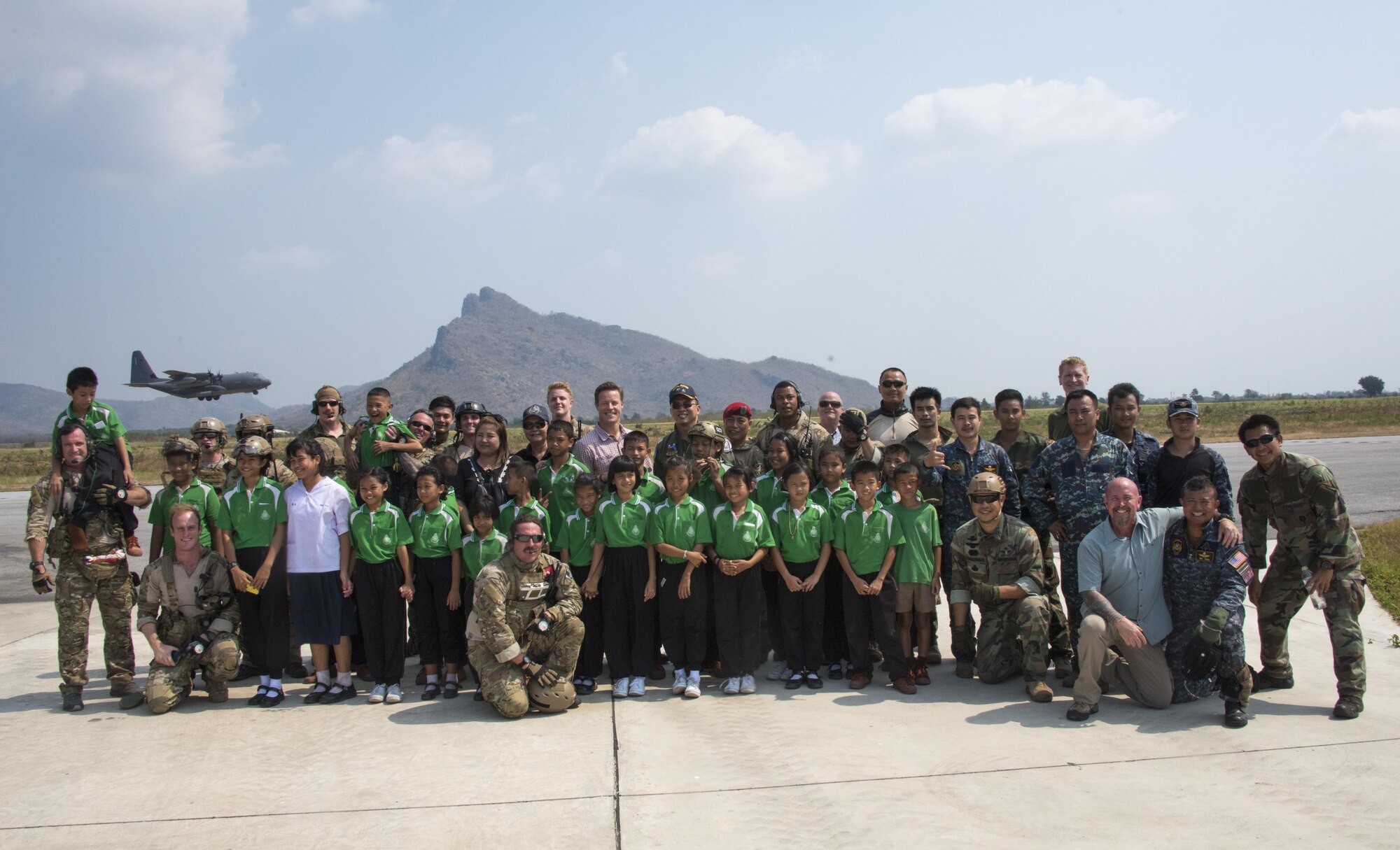 U.S. Air Force MC-130J Commando II assigned to the 17th Special Operations Squadron conducts a low-level flyover as combat controllers and pararesucement from the Royal Thai Air Force Special Operations Regiment and 320th Special Tactics Squadron pose for a group photo with Thai students from Sirijantaranmit School Lopburi, Feb. 23, 2017 at Thai Air Force Base Wing 2 in Lopburi, Thailand. Military members concluded Cobra Gold 2017 with a community relations event which included a $4,000 donation of computers and school supplies to a local Thai school. (U.S. Air Force photo by Capt. Jessica Tait)