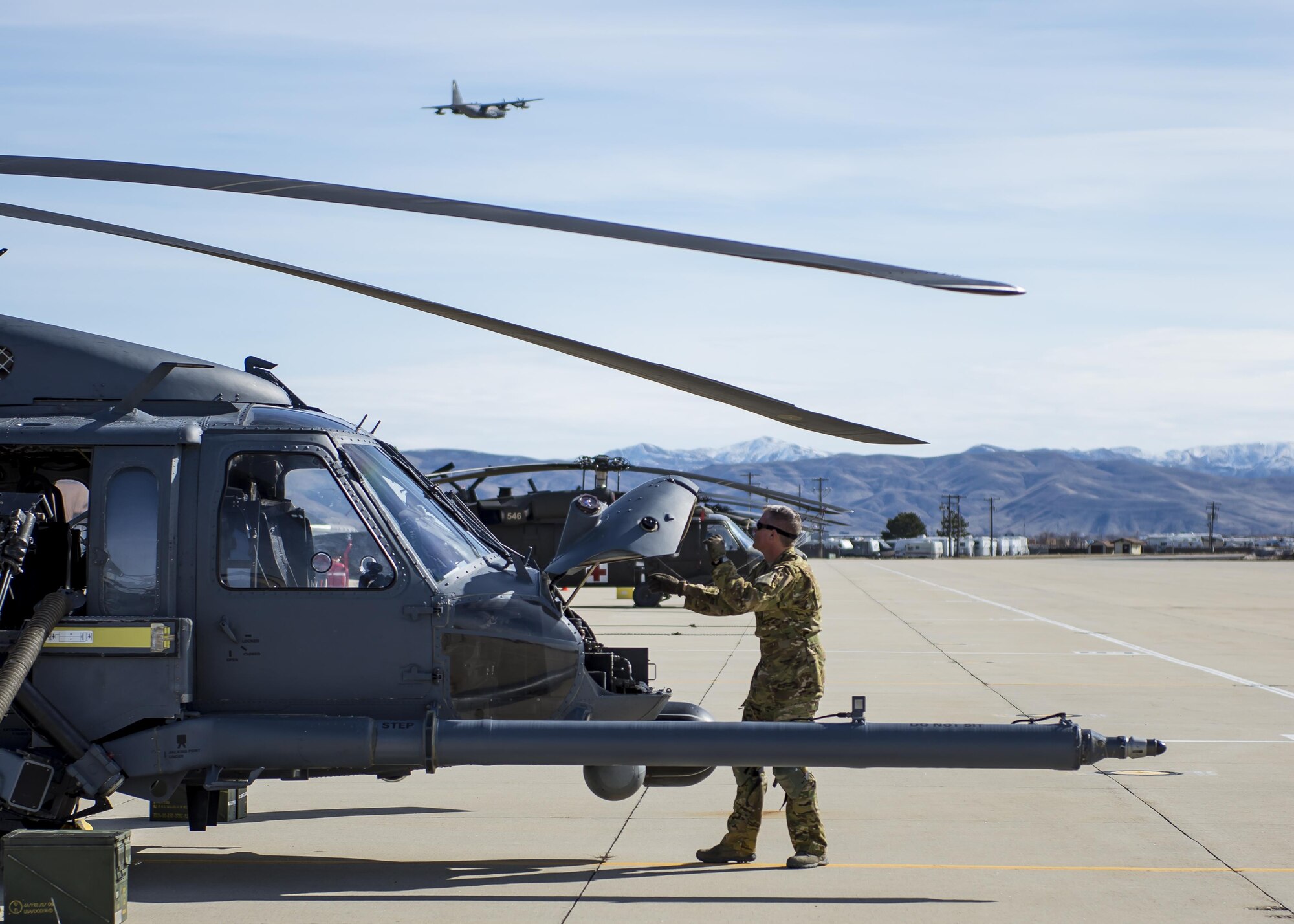 An Airmen with the 943rd Rescue Group performs a pre-flight inspection on an HH-60G Pave Hawk helicopter as HC-130J Combat King II flies over head, March 3, 2017, at Gowen Field, Idaho, during pre-deployment training for the 305th Rescue Squadron. The 305th RQS, located at Davis-Monthan Air Force Base, Ariz., is conducting training at the Idaho Air National Guard’s Orchard Combat Training Center, a 143,000-acre live-fire range located south of Boise, Idaho, to hone all of their search and rescue procedures before heading to Southwest Asia. (U.S. Air Force photo/Staff Sgt. Jared Trimarchi)