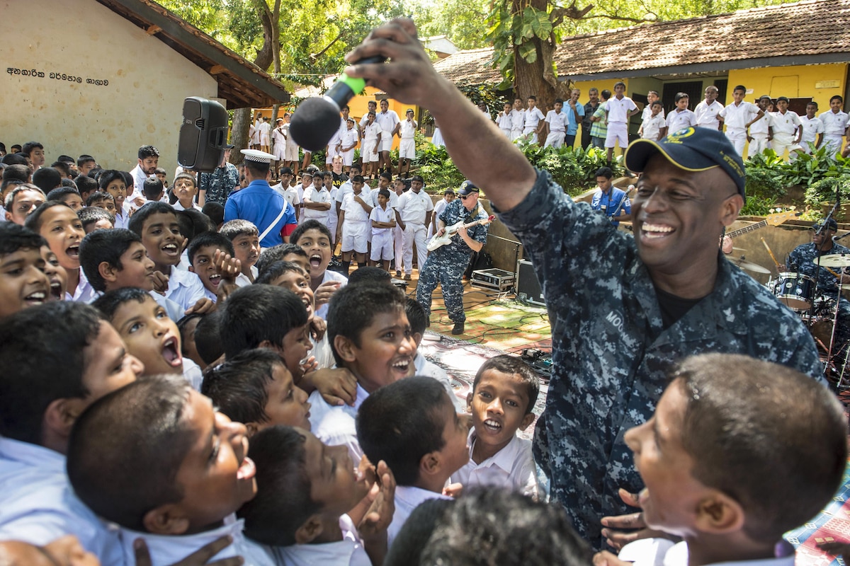 A sailor sings to children who surround him and smile.