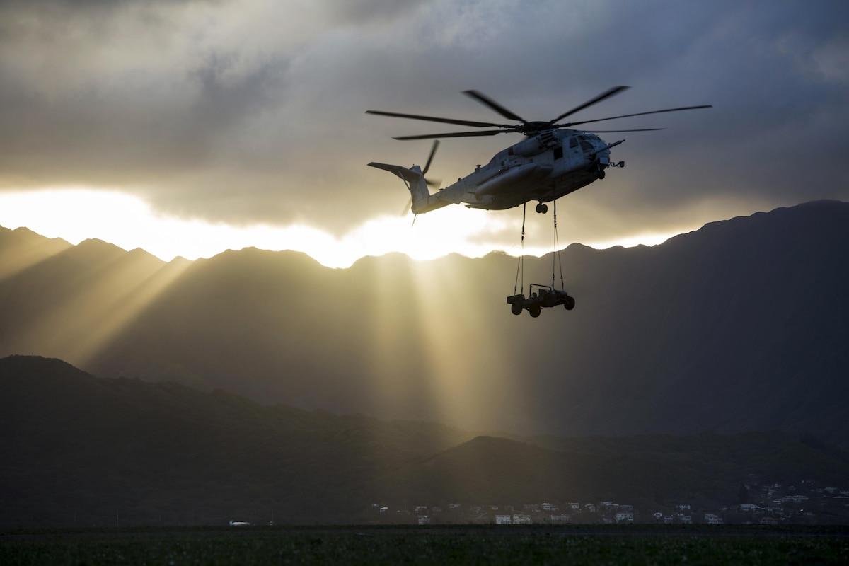 A helicopter carries a Humvee as the sun's rays spread over a mountain range.
