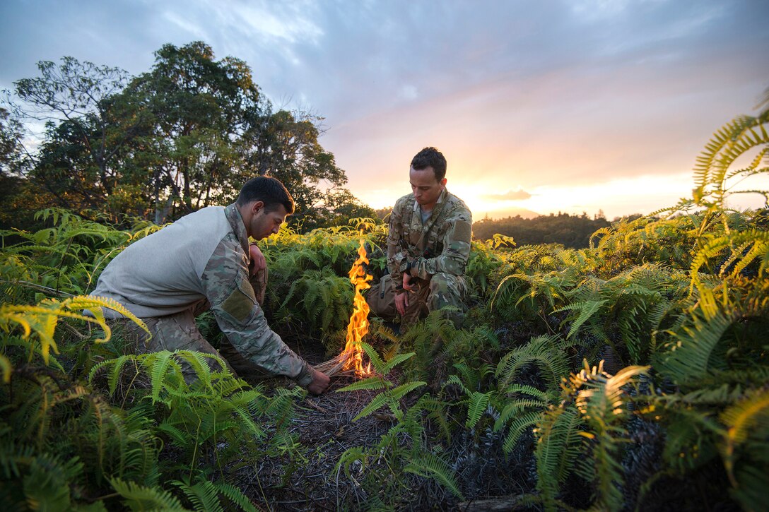 Air Force Senior Airmen Alex Triani, left, adds wood to a fire while Jonathan Harvey, right, looks on during survival training at the Army's Jungle Operations Training Course in Hawaii, March 7, 2017. Harvey and Triani are survival, evasion, resistance and escape specialist assigned to the 106th Rescue Wing. Air National Guard photo by Staff Sgt. Christopher S. Muncy

