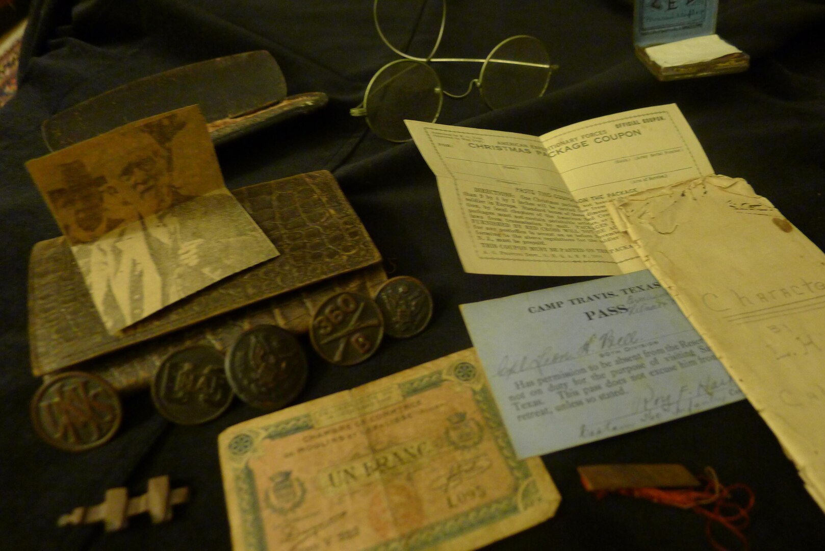From left: French emergency currency; unidentified uniform insignia; Army uniform buttons, wallet; spectacles and case; newspaper photo of unidentified man, French cigarette rolling papers; label for Christmas package; liberty pass from Camp Travis, San Antonio; remains of medal or pin; 1913 essay on character.