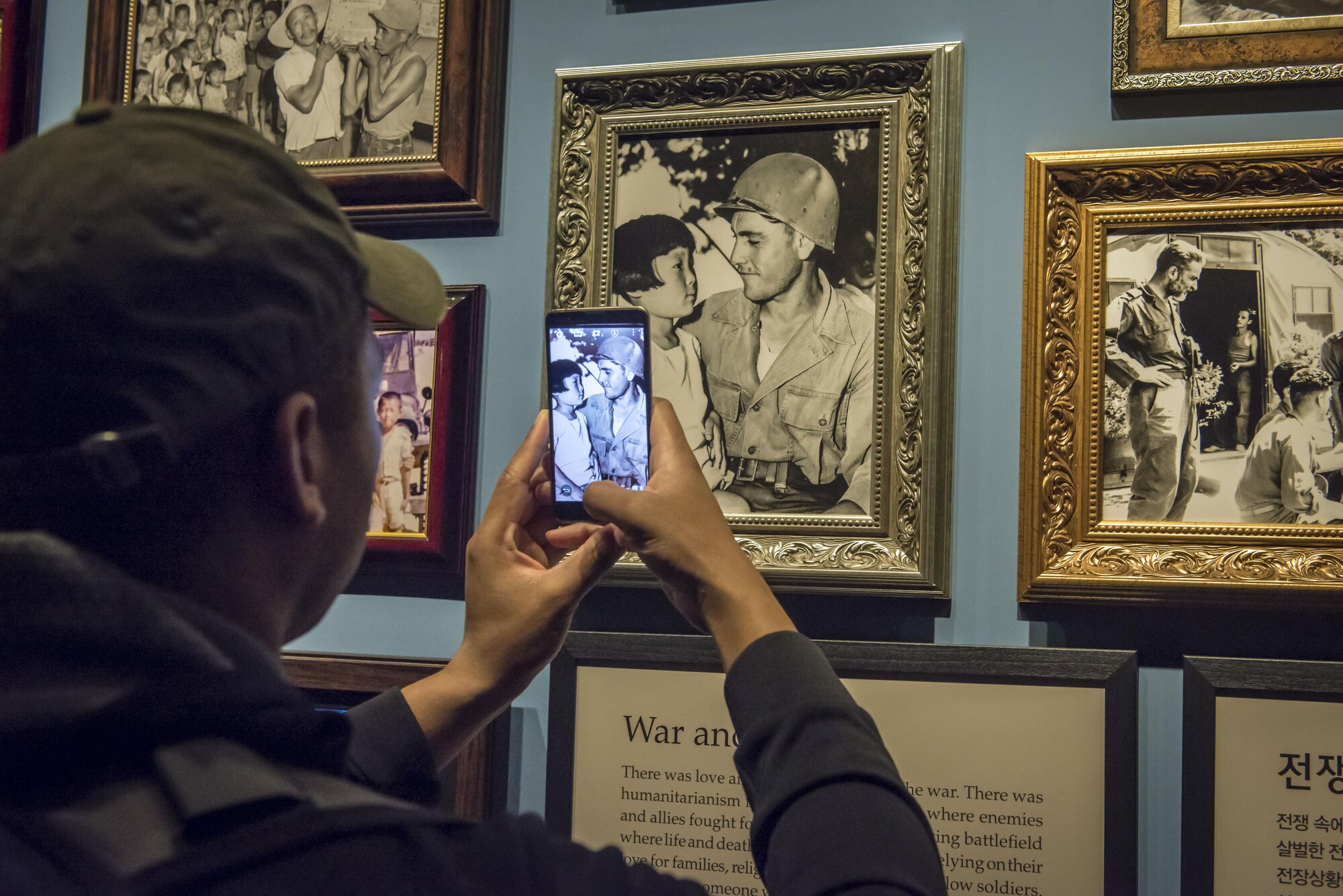 U.S. Air Force Tech. Sgt. Malo Jones , an administration craftsman home stationed at  Headquarters Air Force at the Pentagon in the District of Columbia, snaps a photo with his smartphone of a U.S. soldier and a North Korean girl framed in time by the black and white portrait from the Korean War at the War Memorial of Korea in Seoul, Republic of Korea, March 18, 2017. A plaque just below this photo entitled, “War and Love,” reads, “There was love among soldiers even in the war. There was humanitarianism in the bloody battlefield where enemies and allies fought for their lives. In the terrifying battlefield where life and death intersected, they fought relying on their love for families, religious beliefs and trust of fellow soldiers. And when someone was injured, regardless of whether the person was friend or foe, as human beings they took care of the person. (U.S. Air Force photo by Staff Sgt. Benjamin W. Stratton)