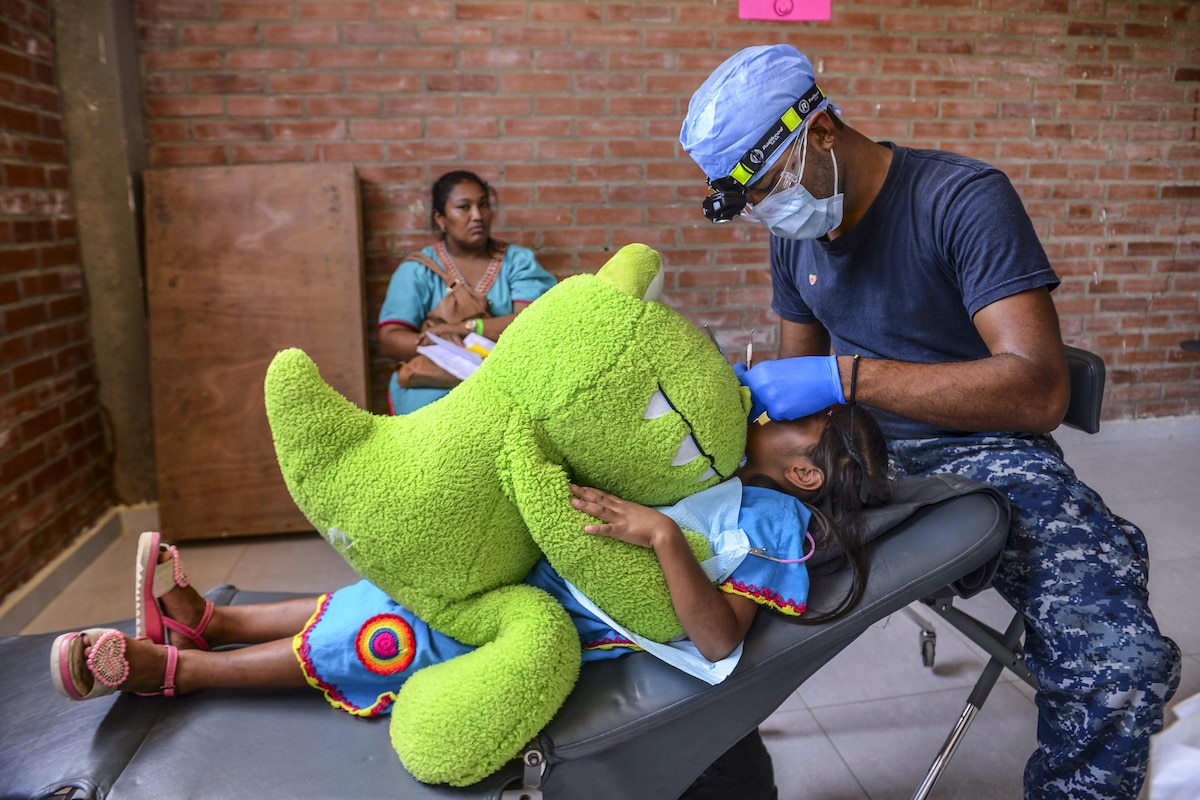 A sailor cleans a child’s teeth as she holds a large stuffed animal.