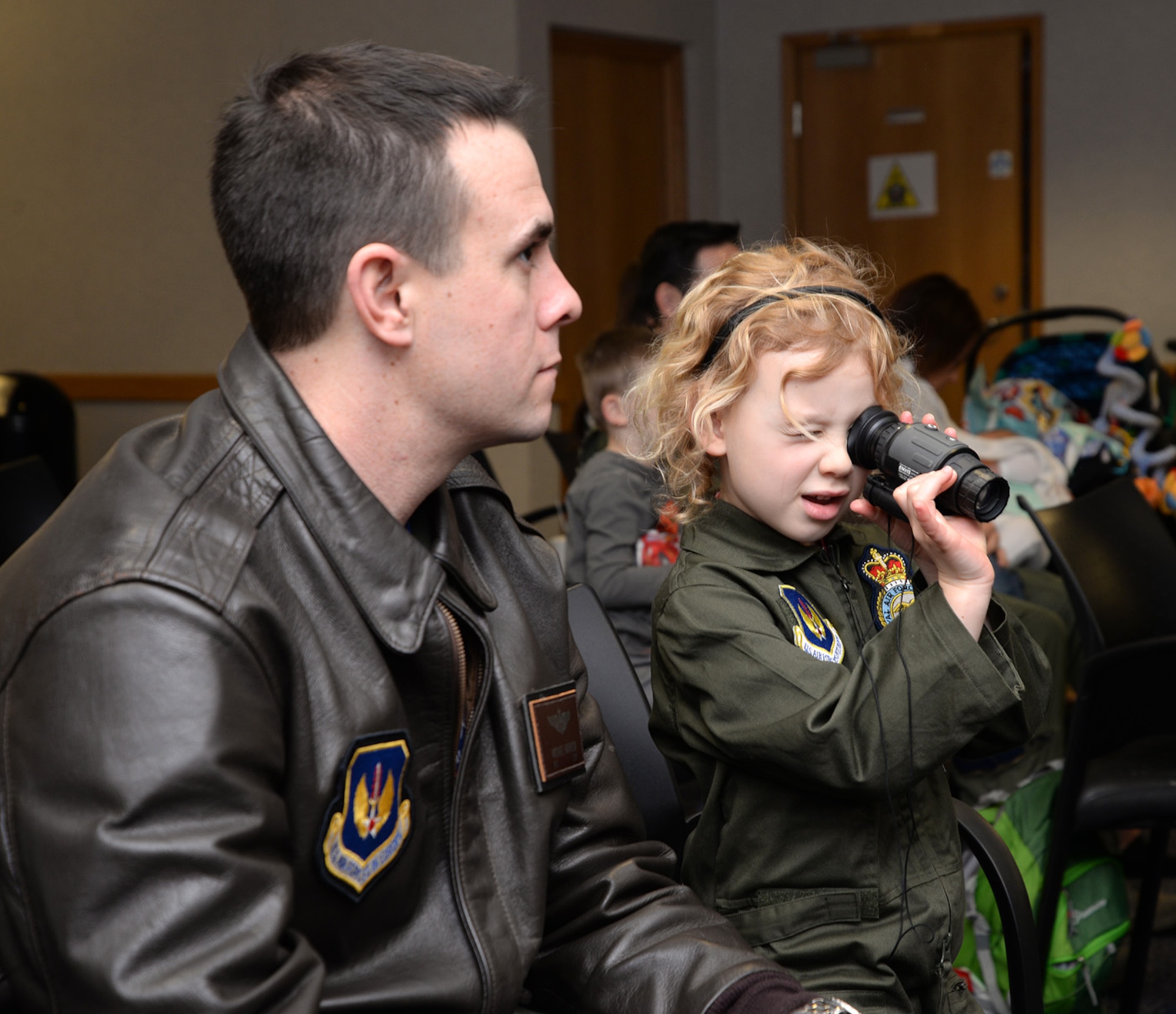 U.S. Air Force Maj. Michael Harrison, left, 100th Operations Support Squadron assistant director of operations, listens to a survival, evasion, resistance and escape instructor as his daughter Isabella, 5, checks out a night-vision monocular at the 100th Operations Group “Bring Your Child to Work Day” as part of Month of the Military Child April 28, 2017, on RAF Mildenhall, England. During the kid-friendly SERE training, children tried on equipment including helmets, and had their faces painted with camouflage pattern. (U.S. Air Force photo by Karen Abeyasekere) 