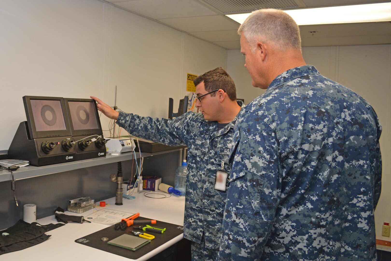 Petty Officer 1st Class Andrew Leigh (left) demonstrates to Rear Adm. Jon Kreitz how Sailors at Southeast Regional Maintenance Center (SERMC) test, clean and repair fiber optic cable. The 2M (Miniature/Micro-miniature) shop at SERMC can inspect, repair, fabricate and troubleshoot fiber optic cable onboard all applicable classes of U.S. Navy surface ships. This image was manipulated using filters to blur portions for security and/or privacy concerns. 