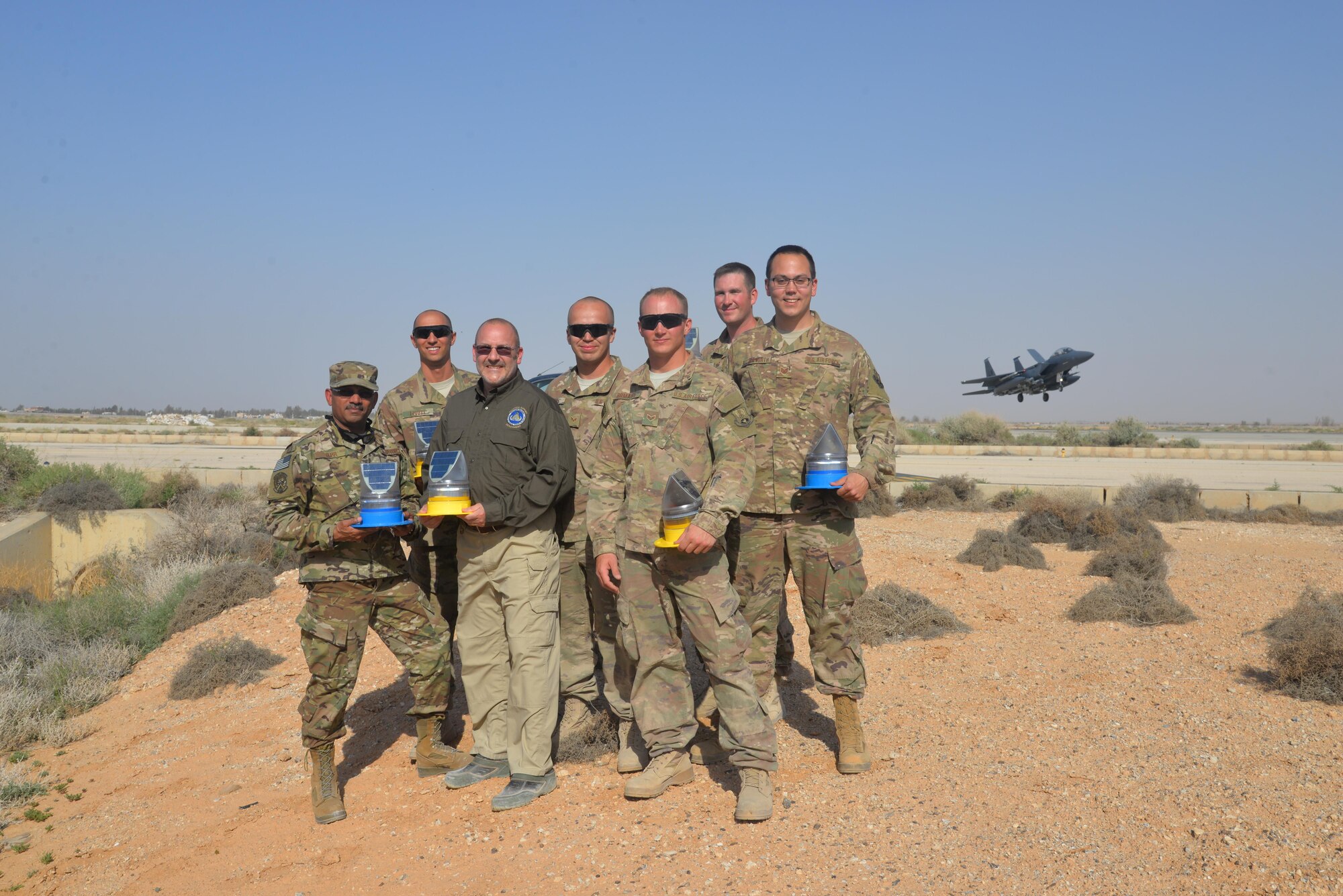Members from the 332nd Expeditionary Civil Engineer Squadron pose with solar lights as an F-15E Strike Eagle lands behind them, May 10th, 2017, in Southwest Asia.  The 332nd ECES squadron installed solar lights on the flight line, making it safer for pilots during night operations. (U.S. Air Force Photo by Staff Sgt. Samuel O’Brien)