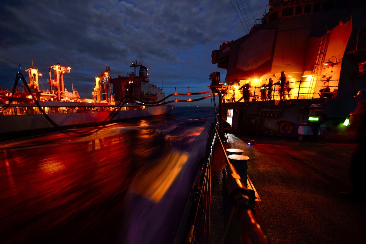 Two ships conduct a night replenishment at sea as yellow lights flare.
