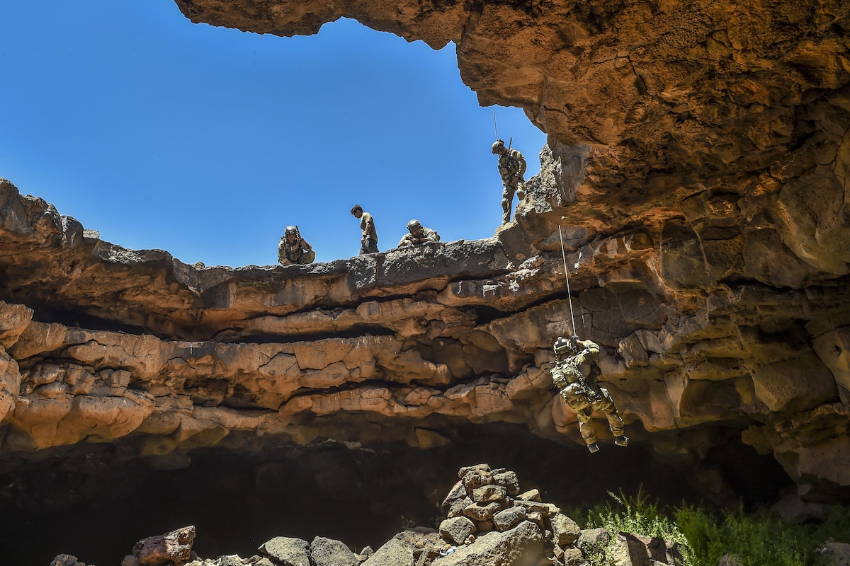 An airman rappels into a cave complex with blue skies overhead.