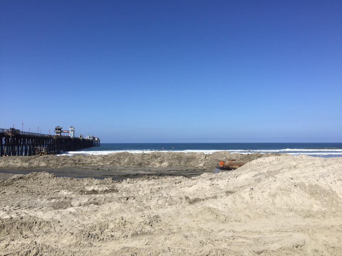 Material dredged from the annual Oceanside Harbor channel maintenance project lies adjacent to the Oceanside Pier. As the sun bleaches the sand, it takes on the appearance of material along the shoreline. Construction equipment then contours the sand to increase beach width and enhance protection from winter storms.