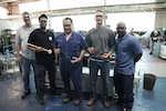 Members of the Norfolk Naval Shipyard Rapid Prototype Lab and Shop 72 (Rigging Department) Lifting and Handling Specialist Jonathan Woodruff showcase the progression of prototypes of the Tie Bolt Anti-Rotation Tool. 