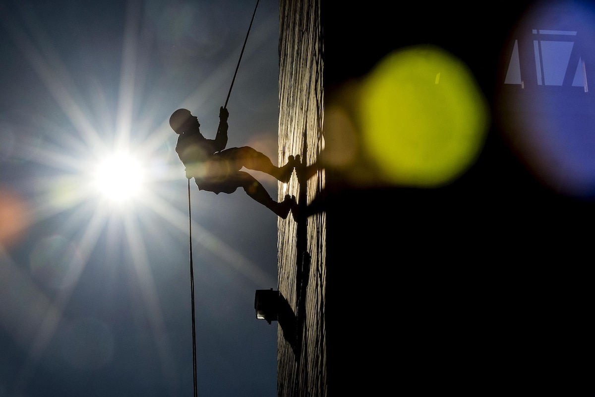 A soldier rappels down a wall at night with a bright light shining.